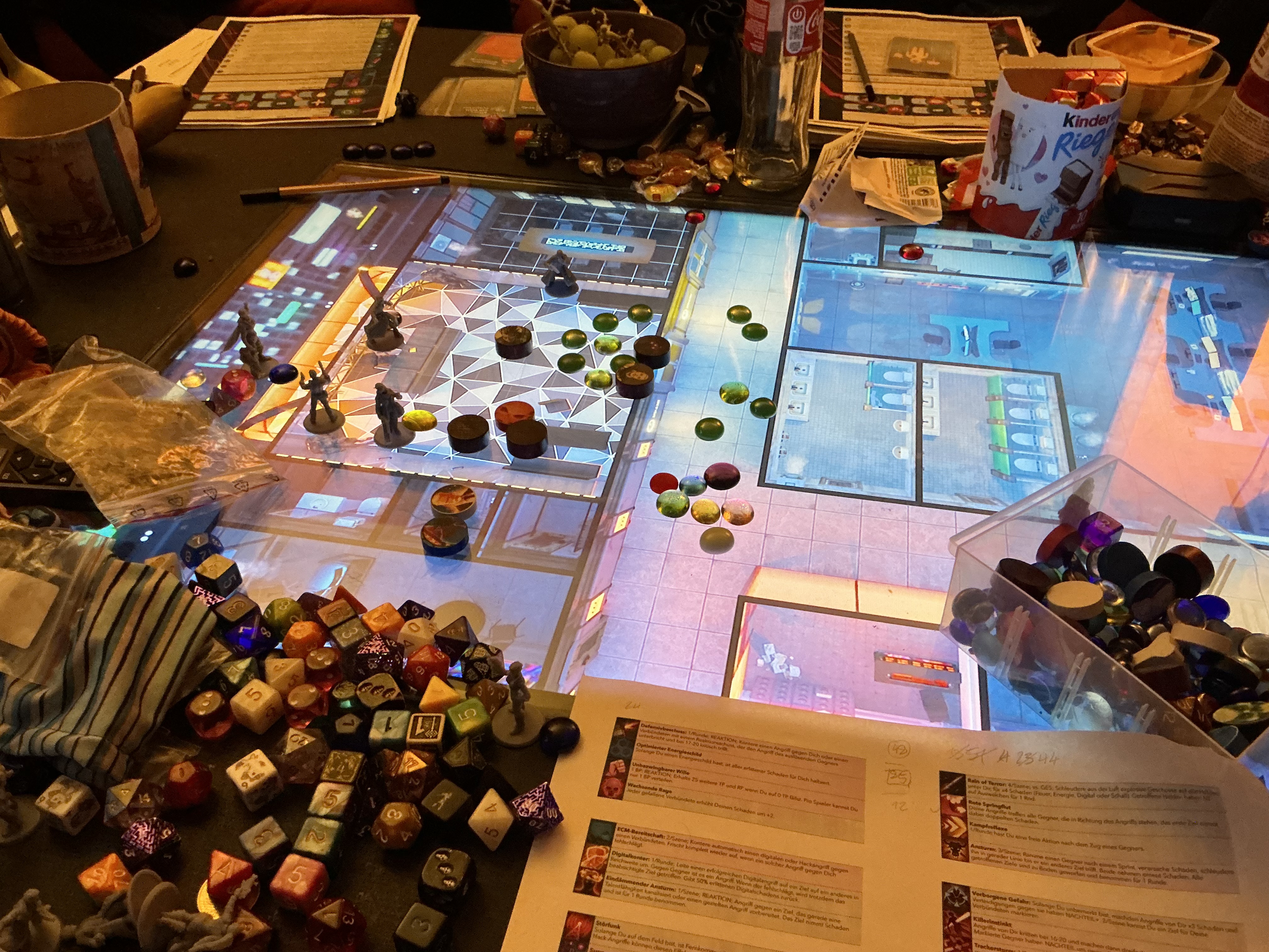 Picture of the gaming table with the animated map of an office floor with miniatures and tokens showing the position of characters in the scene. Lots of dice, GM notes, character sheets and snacks are scattered around it.