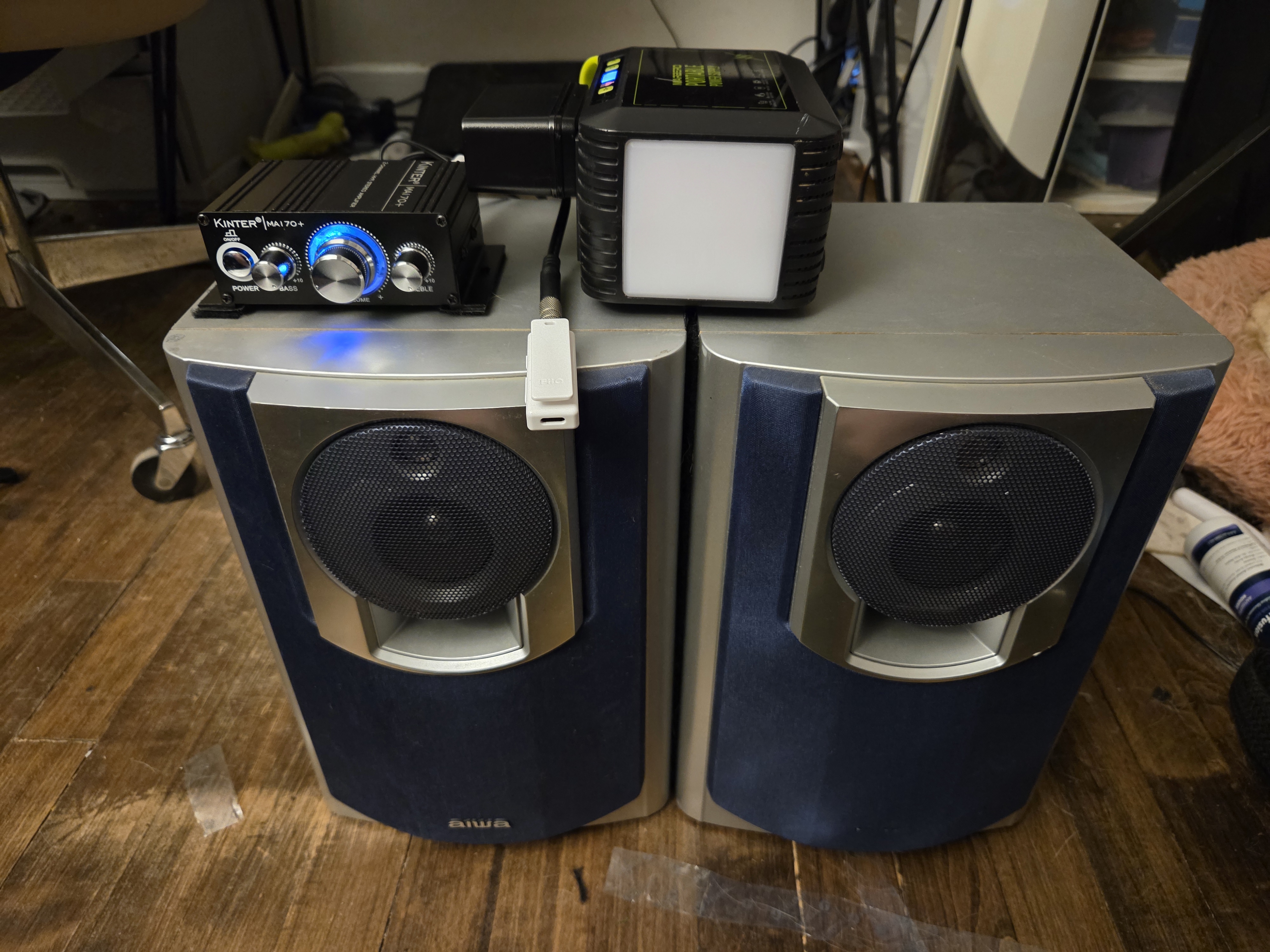 A set of bookshelf speakers velcro'd to each other with a small speaker amp velcro'd to the left speaker, there's a Bluetooth dongle and a battery bank sitting next to it